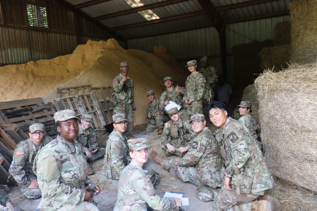 A group of Army ROTC students in camouflage military uniforms gather inside a barn with metal walls and piles of hay around them. Some sit or kneel on the ground while others stand, listening and holding papers. Several smile at the camera and pose for the photo. 