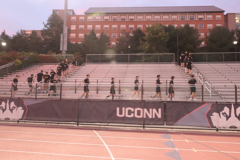 A group of Army ROTC students wearing black shirts with yellow ‘ARMY’ lettering exercise on metal bleachers at the University of Connecticut stadium. They run up and down the steps while a red running track and a banner reading ‘UCONN’ with Husky logos are visible on the fence in front. 